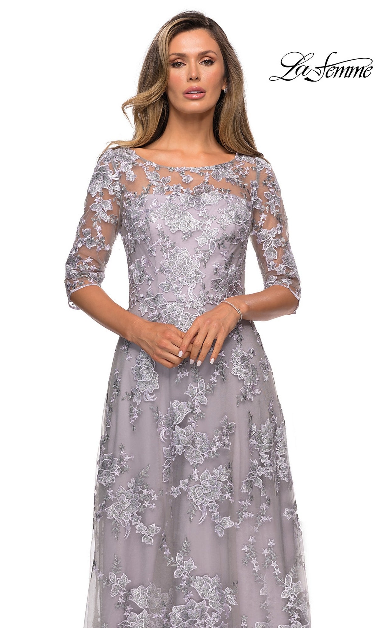 Woman modeling the Long Formal Dress: La Femme 27854 in lavender lace with floral embroidery and sheer sleeves, set against a white background. “La Femme” logo at top right.